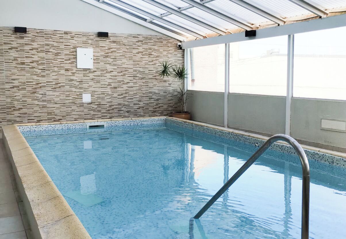 En indendørs swimmingpool med fliserede vægge, en metalstige og en potteplante.