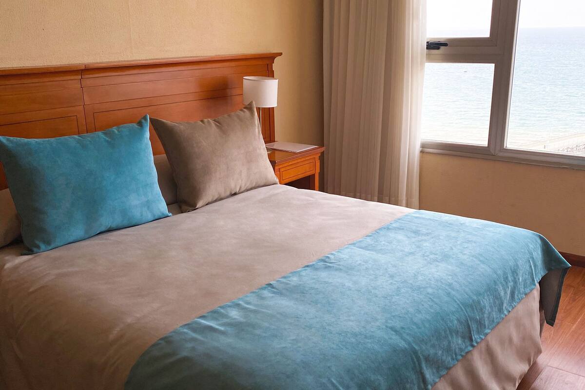 Un lit avec une tête de lit en bois, une table de chevet avec une lampe et une vue sur la mer par la fenêtre.