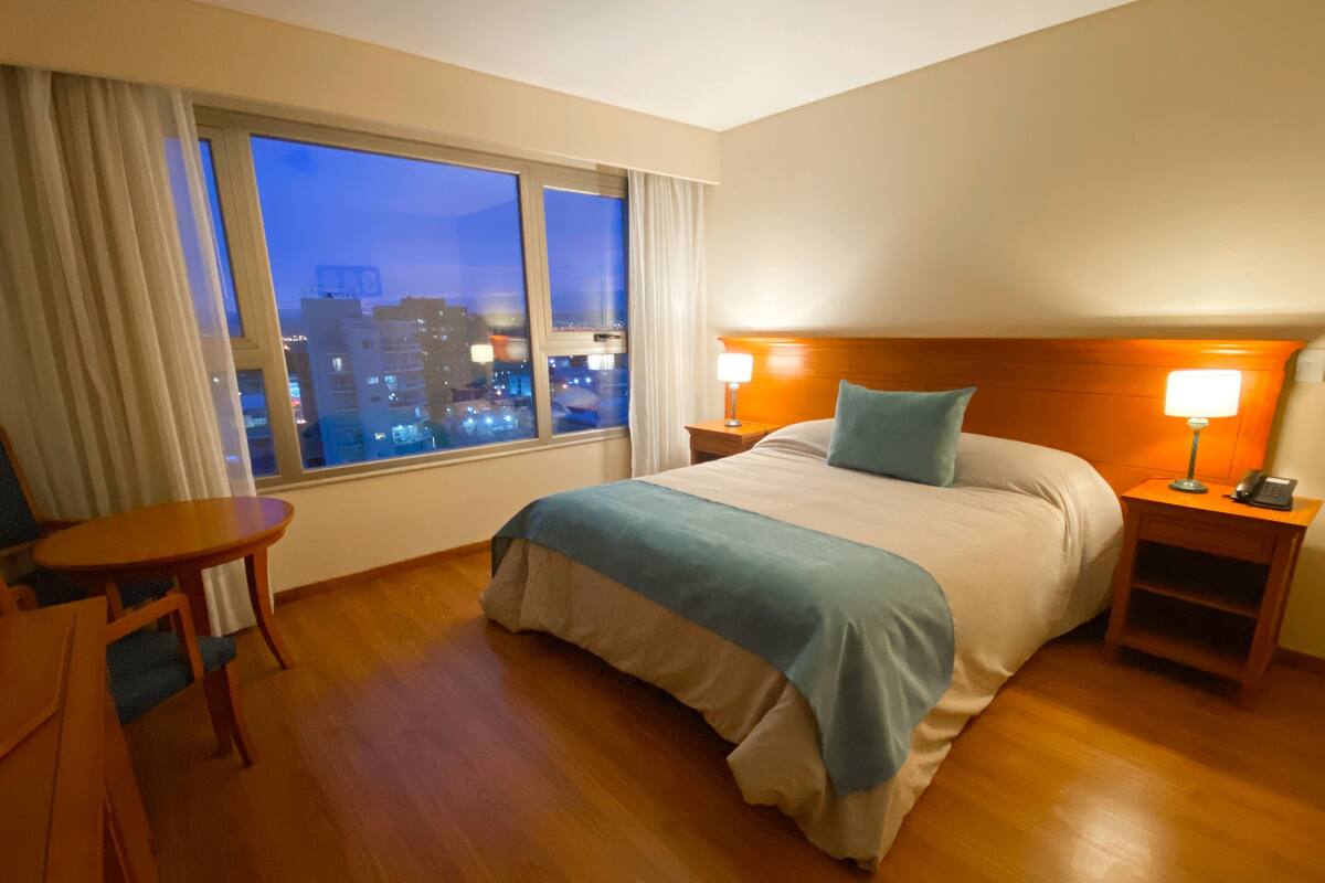Une chambre d’hôtel avec un grand lit, deux tables de chevet avec des lampes, un fauteuil et une vue sur la ville au crépuscule.