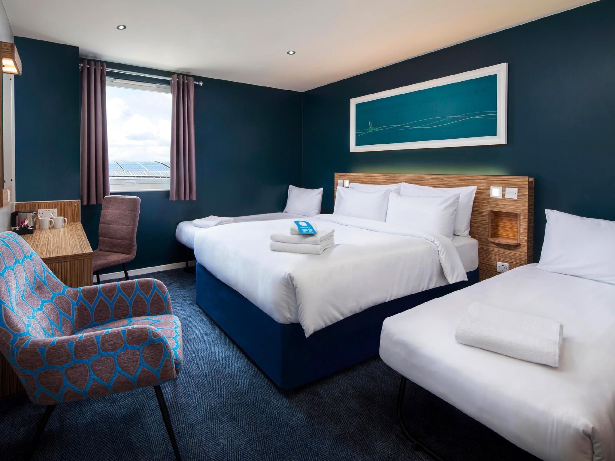 Une chambre d’hôtel avec un grand lit, un plus petit lit, une chaise, un bureau et une fenêtre avec des rideaux.