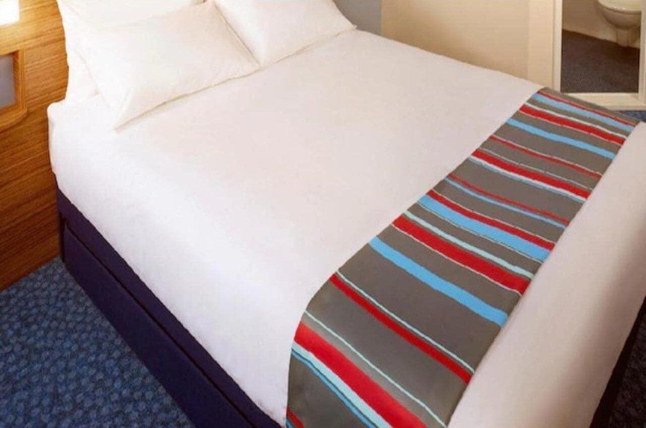 A bed with white linens and a striped blanket, a wooden headboard, and a blue carpeted floor.