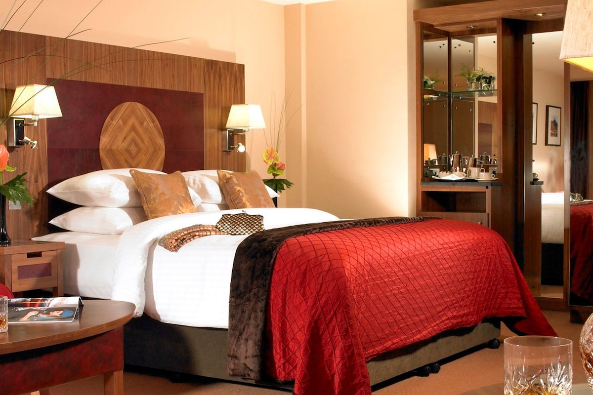 Une chambre d’hôtel avec un grand lit, deux lampes de chevet, une tête de lit en bois, une table de chevet, une table en verre et un miroir.