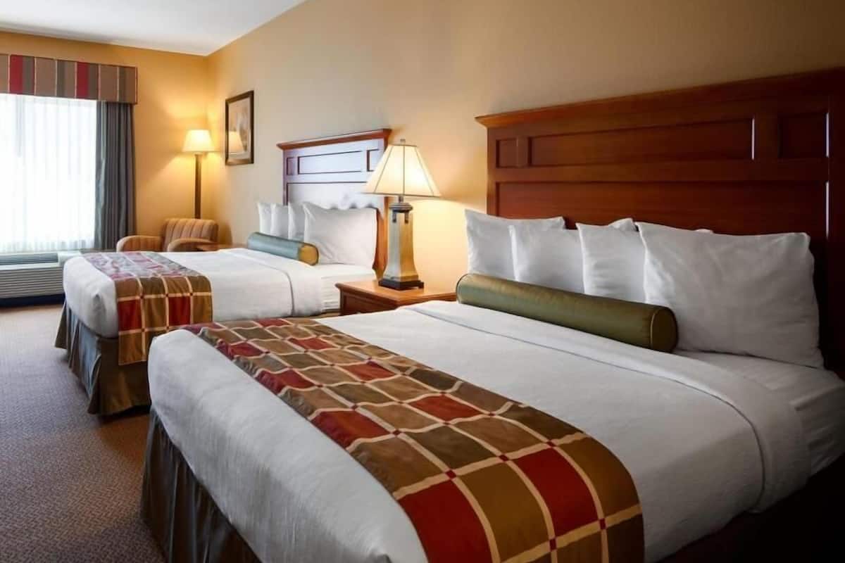 A hotel room with two beds, a nightstand with a lamp, a window with curtains, and a wall-mounted picture.