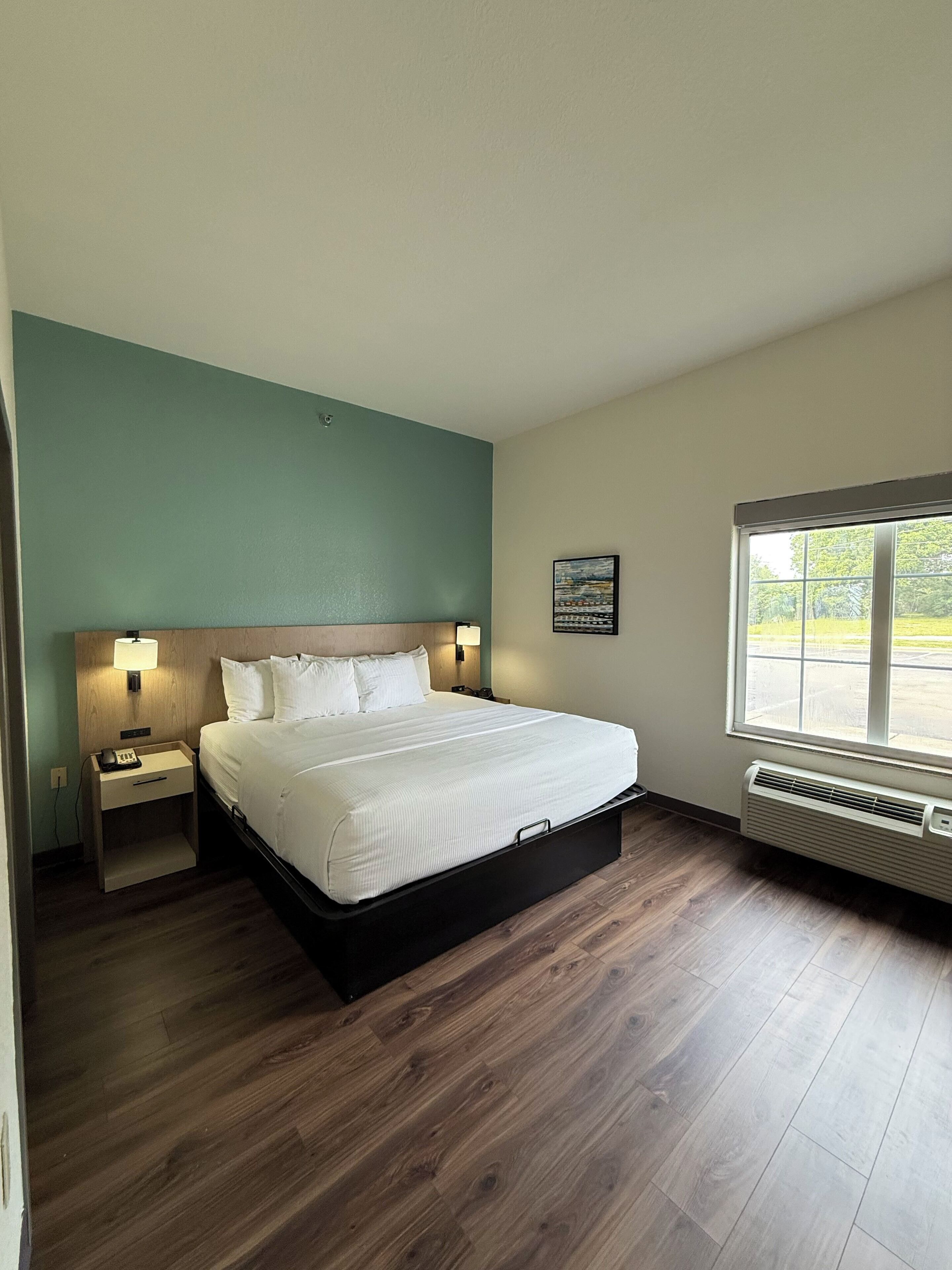 A hotel room with a bed, a nightstand, a wall-mounted lamp, a window with a view, and a wall-mounted air conditioner.