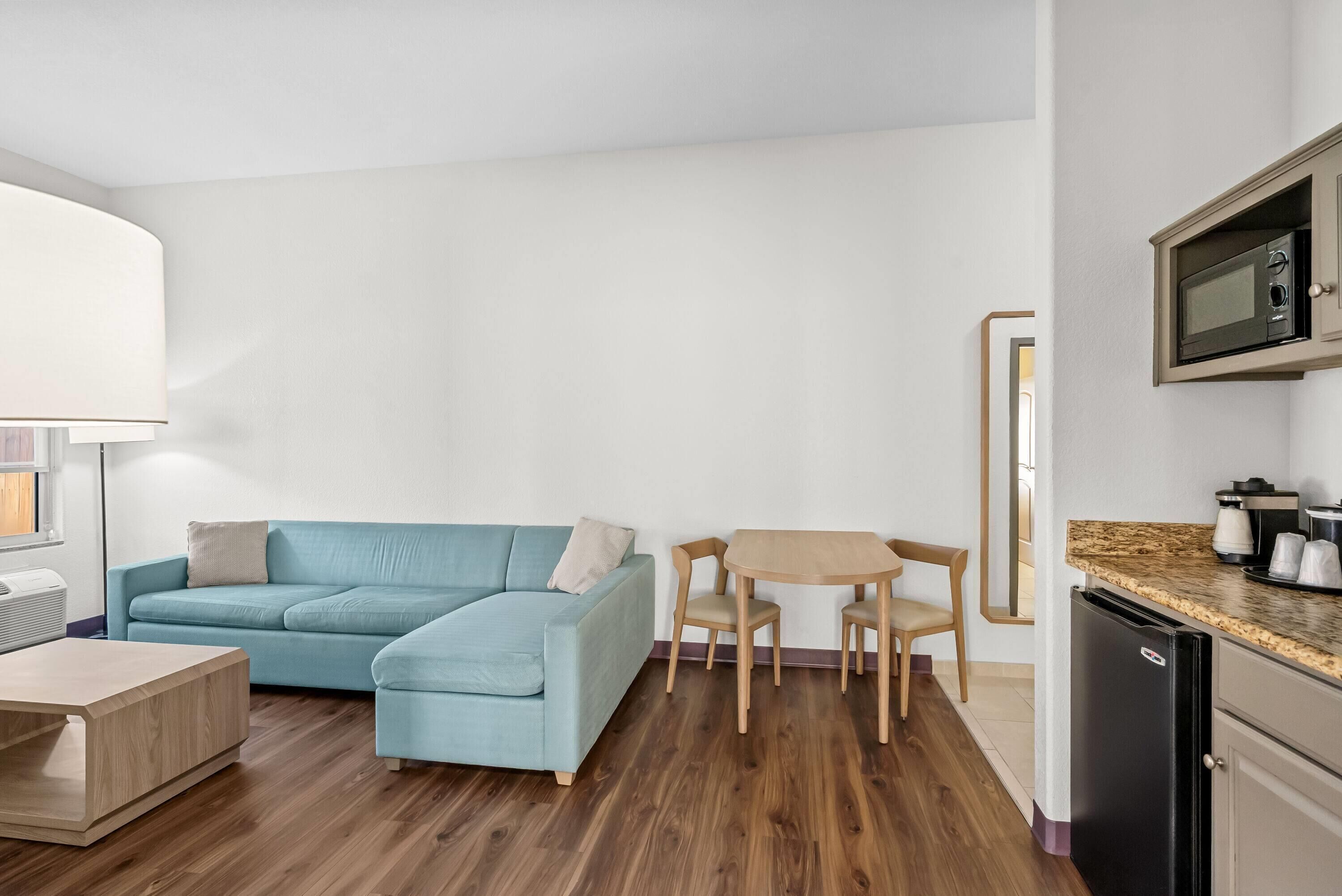A modern living room with a blue sofa, a wooden coffee table, and a small dining table with chairs. A microwave is visible in the kitchen area.