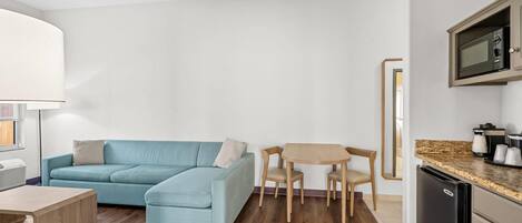 A modern living room with a blue sofa, a wooden coffee table, and a small dining table with chairs. A microwave is visible in the kitchen area.