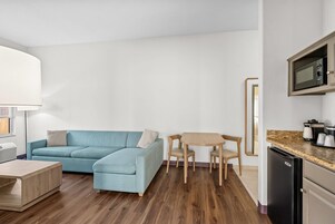 Una sala moderna con un sofá azul, una mesa de centro de madera y una pequeña mesa de comedor con sillas. Se ve un microondas en la zona de la cocina.