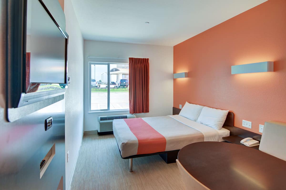 A hotel room with a bed, bedside table, and a view of the parking area.