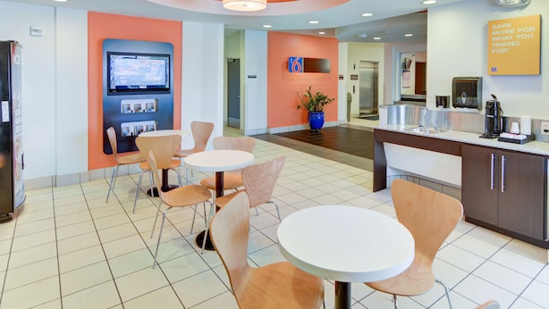 A modern hotel lobby with a seating area, a digital information board, a coffee machine, and a sign encouraging savings for travel. - Motel 6 Roanoke, TX - Northlake - Speedway (Roanoke)