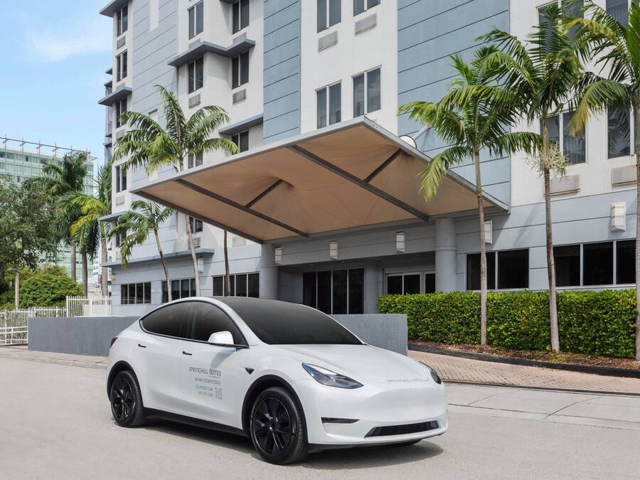 A white Tesla Model 3 parked in front of a modern building with a covered entrance and palm trees.
