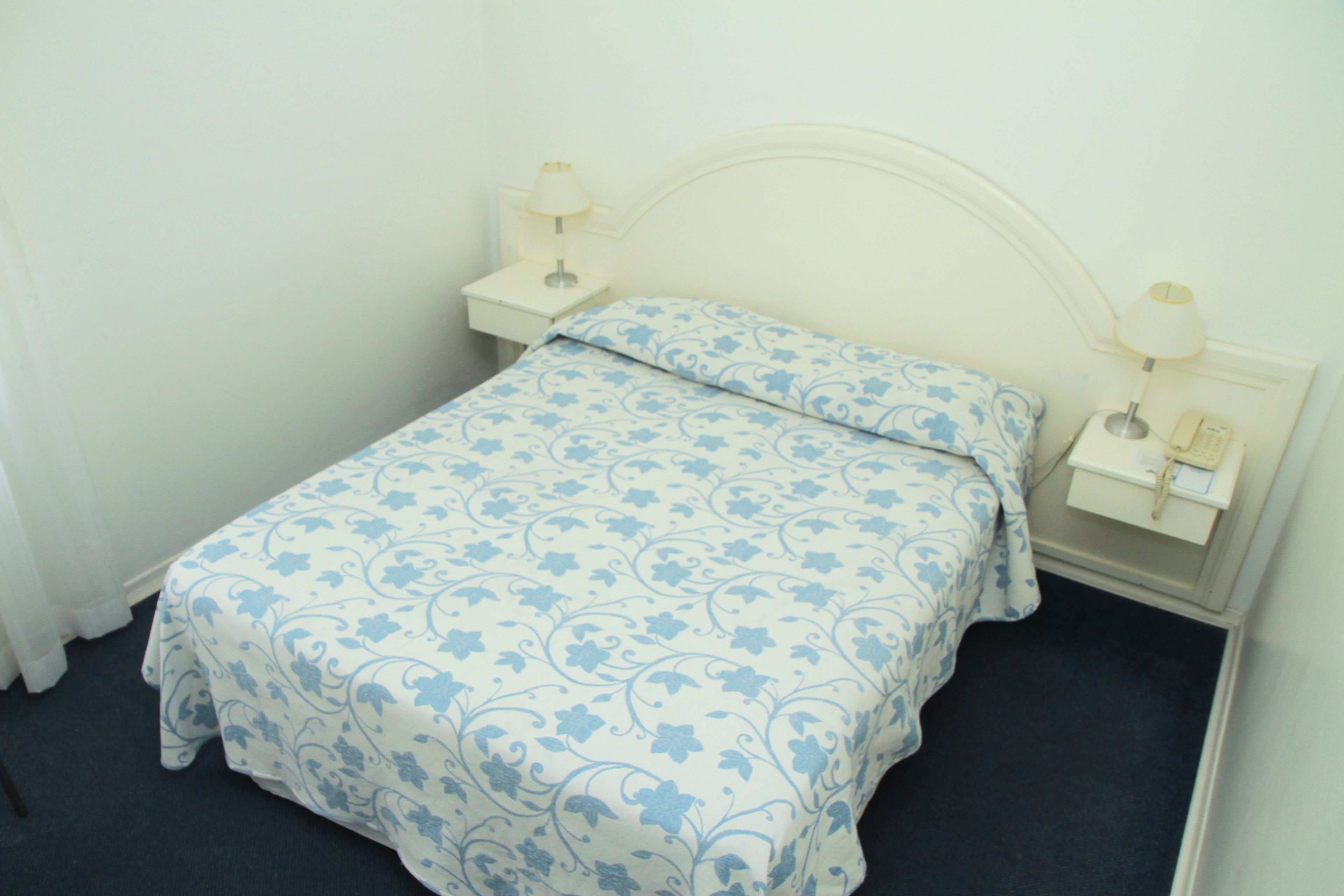 Una cama bien hecha con colcha y almohada estampadas con motivos florales, dos mesitas de noche con lámparas y una mesita auxiliar con un teléfono.