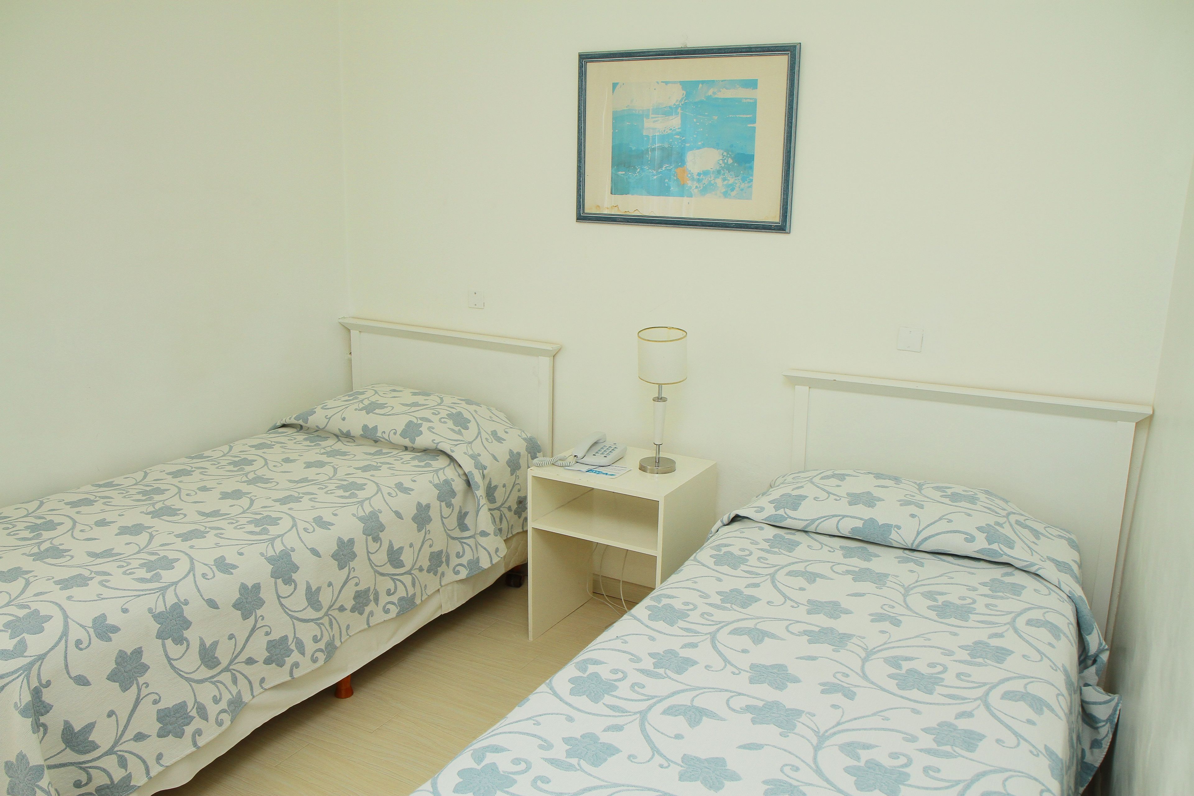 A small, clean room with two single beds, a bedside table with a lamp, and a framed picture on the wall.