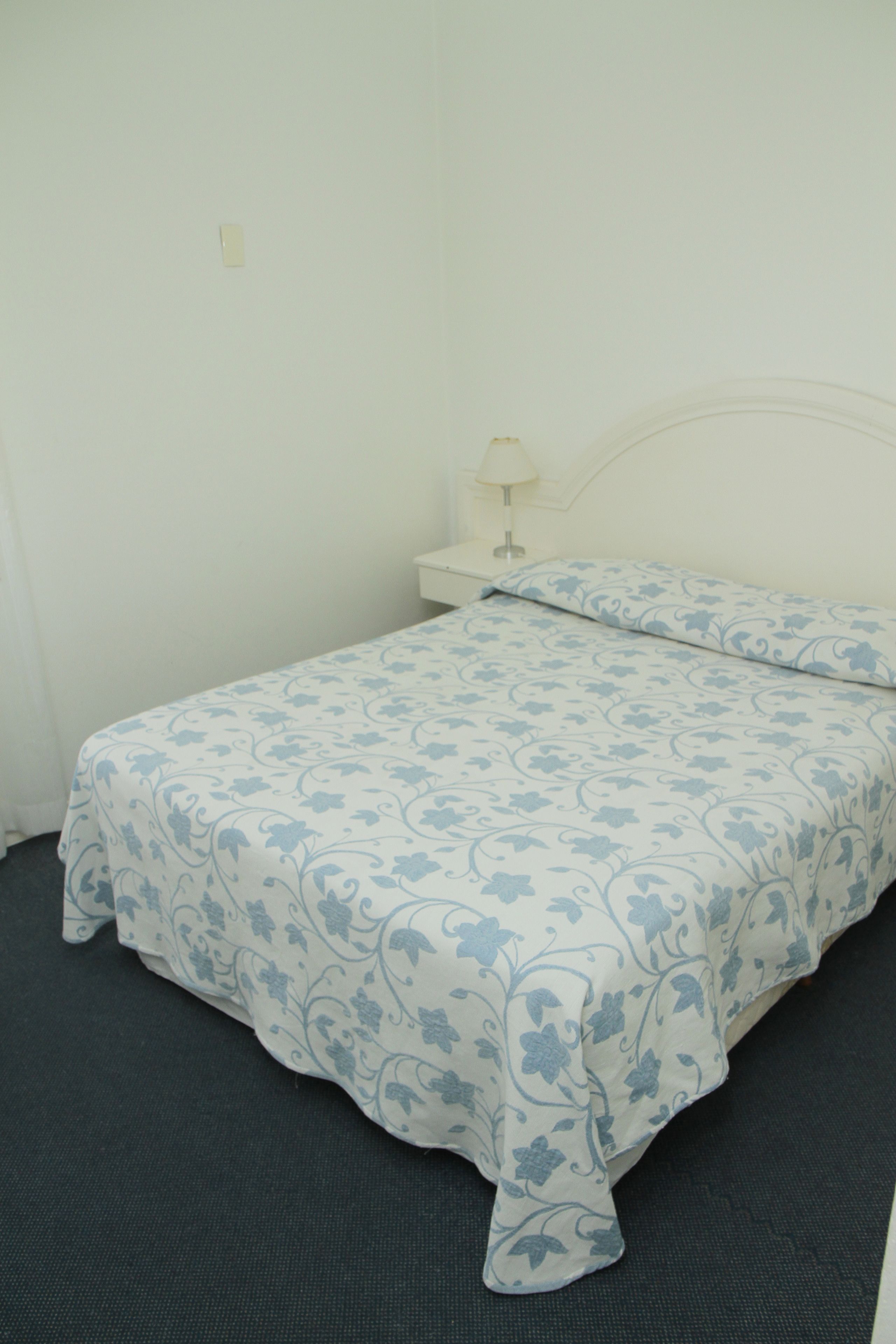 Un llit individual amb un estovat i coixí estampats florals, una taula de nit amb una làmpada, i una paret sense adorns.