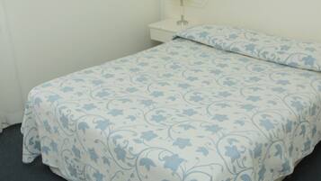 Un llit individual amb un estovat i coixí estampats florals, una taula de nit amb una làmpada, i una paret sense adorns.