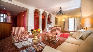A spacious lobby with a red carpet, striped armchairs, a sofa, a glass coffee table with magazines, and a chandelier.