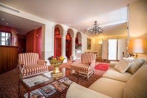 Un hall espacioso con alfombra roja, sillones a rayas, un sofá, una mesa de centro de vidrio con revistas y una araña.