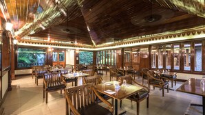 A spacious dining area with wooden tables and chairs, large windows with a view of greenery, and a high wooden ceiling. - Club Mahindra Thekkady (Peermade)
