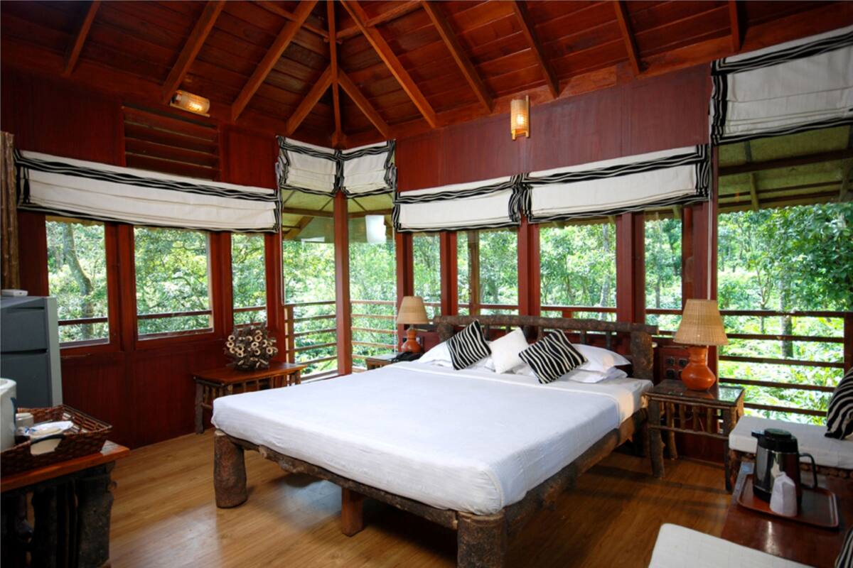 Ein Schlafzimmer in einer Holzhütte mit einem großen Bett, zwei Nachttischen, einem Beistelltisch und einem Fenster mit Blick auf Bäume.
