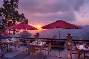 An outdoor dining area with tables and chairs, red umbrellas, and a view of the sunset over a mountainous landscape. - Fort Resorts Mussoorie (Dehradun)