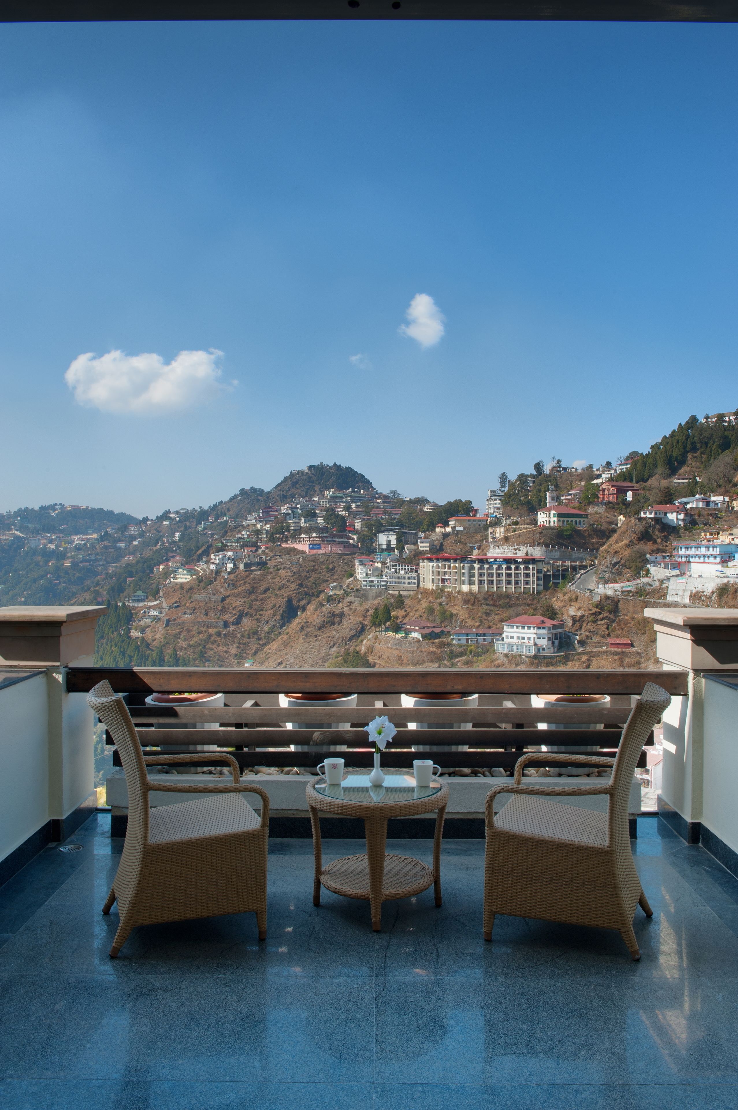A balcony with wicker chairs and a table, overlooking a hillside with buildings.