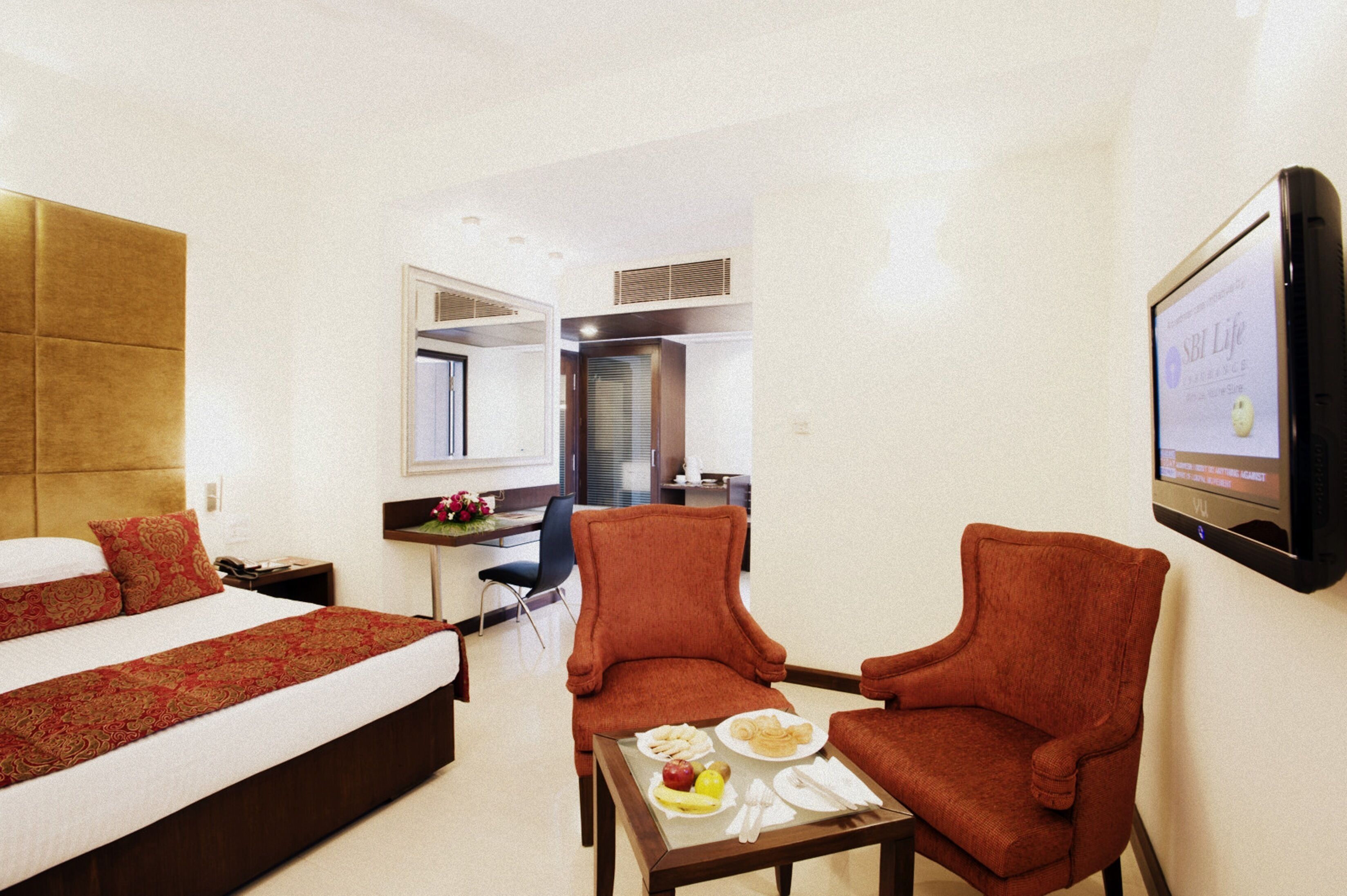 A hotel room with a bed, two armchairs, a small table with breakfast, a TV, and a mirror.