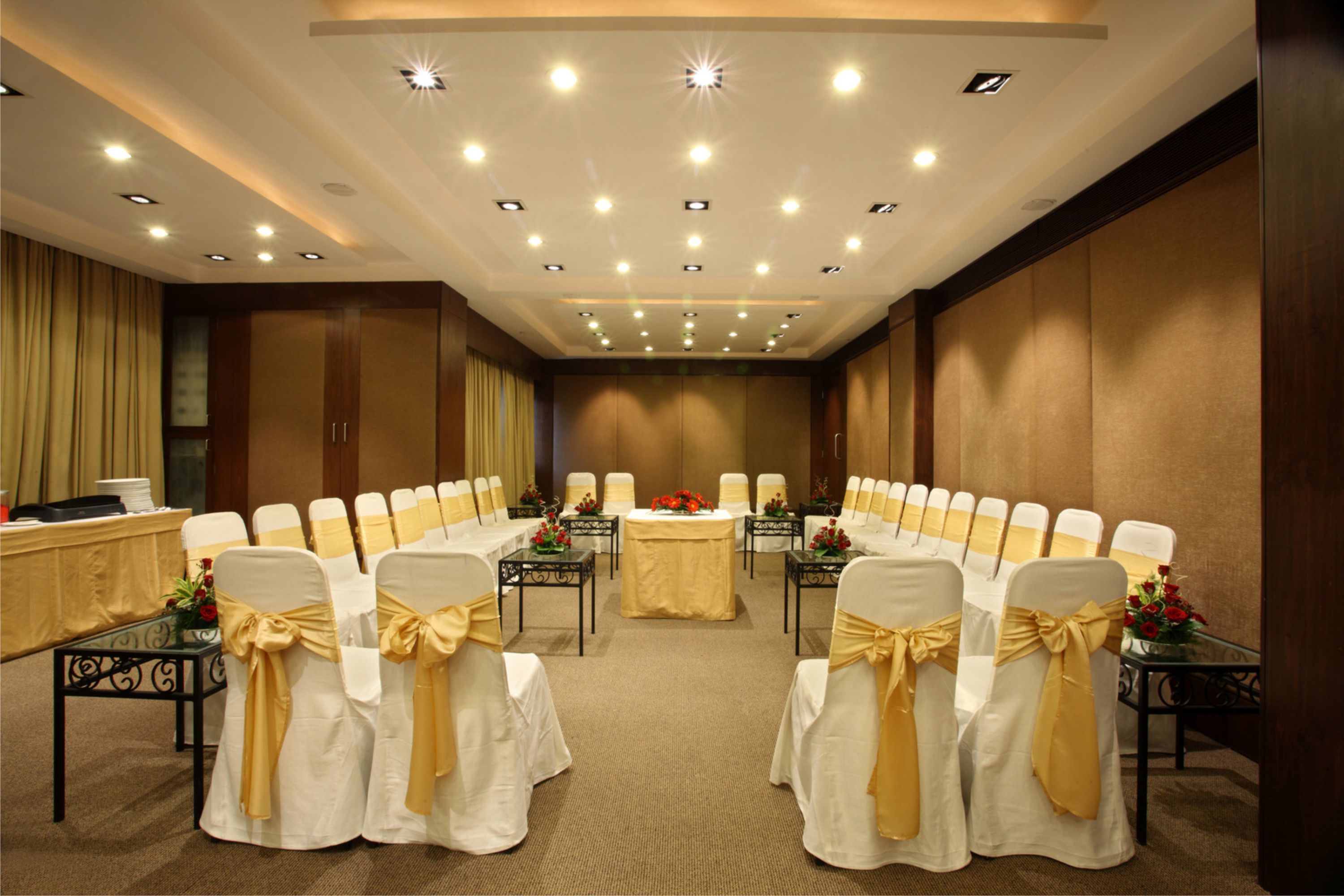 A conference room with rows of white chairs with gold sashes, a central table with a tablecloth, and a ceiling with recessed lighting.