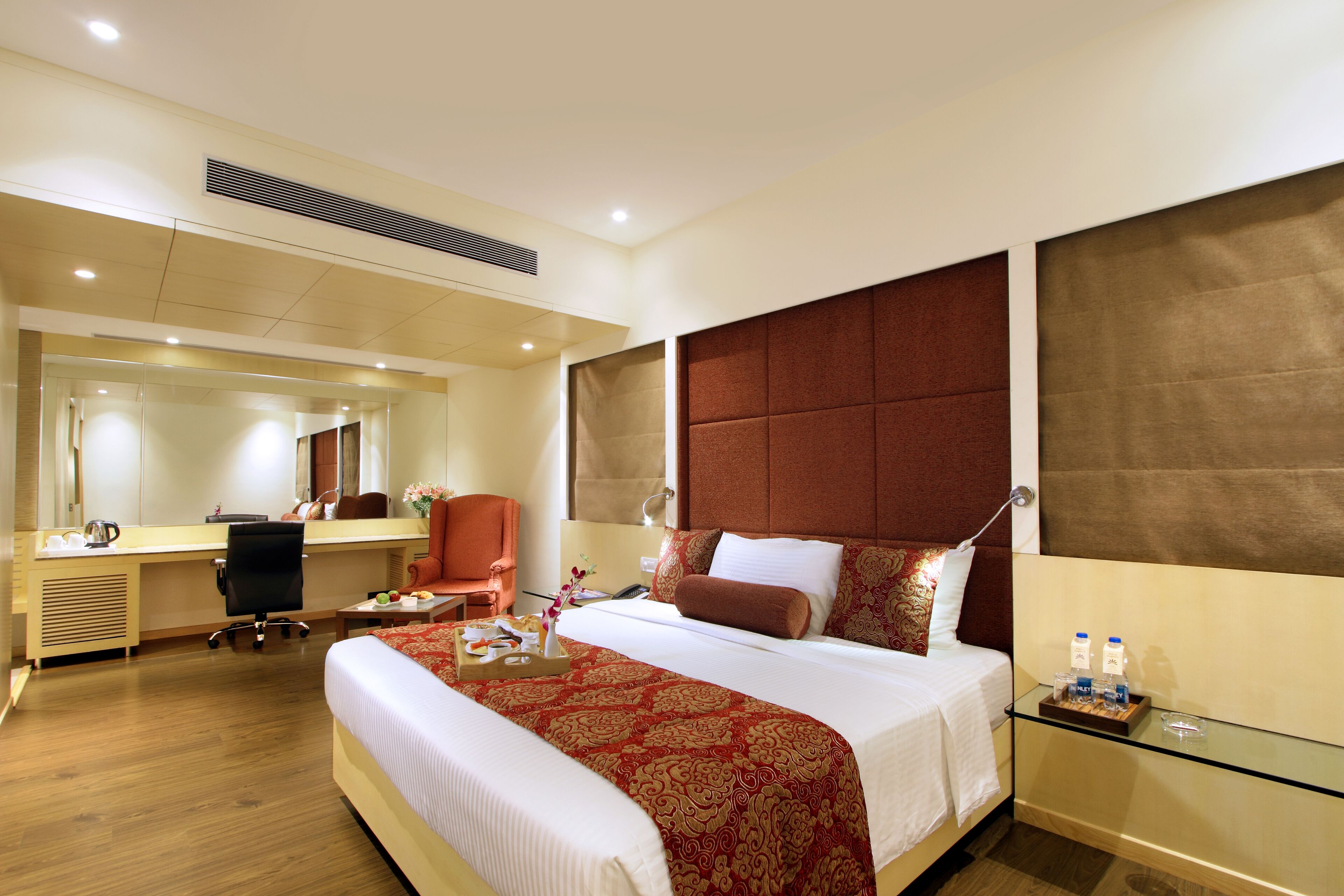 A hotel room with a large bed, a desk, a chair, a bedside table, and a mirror.