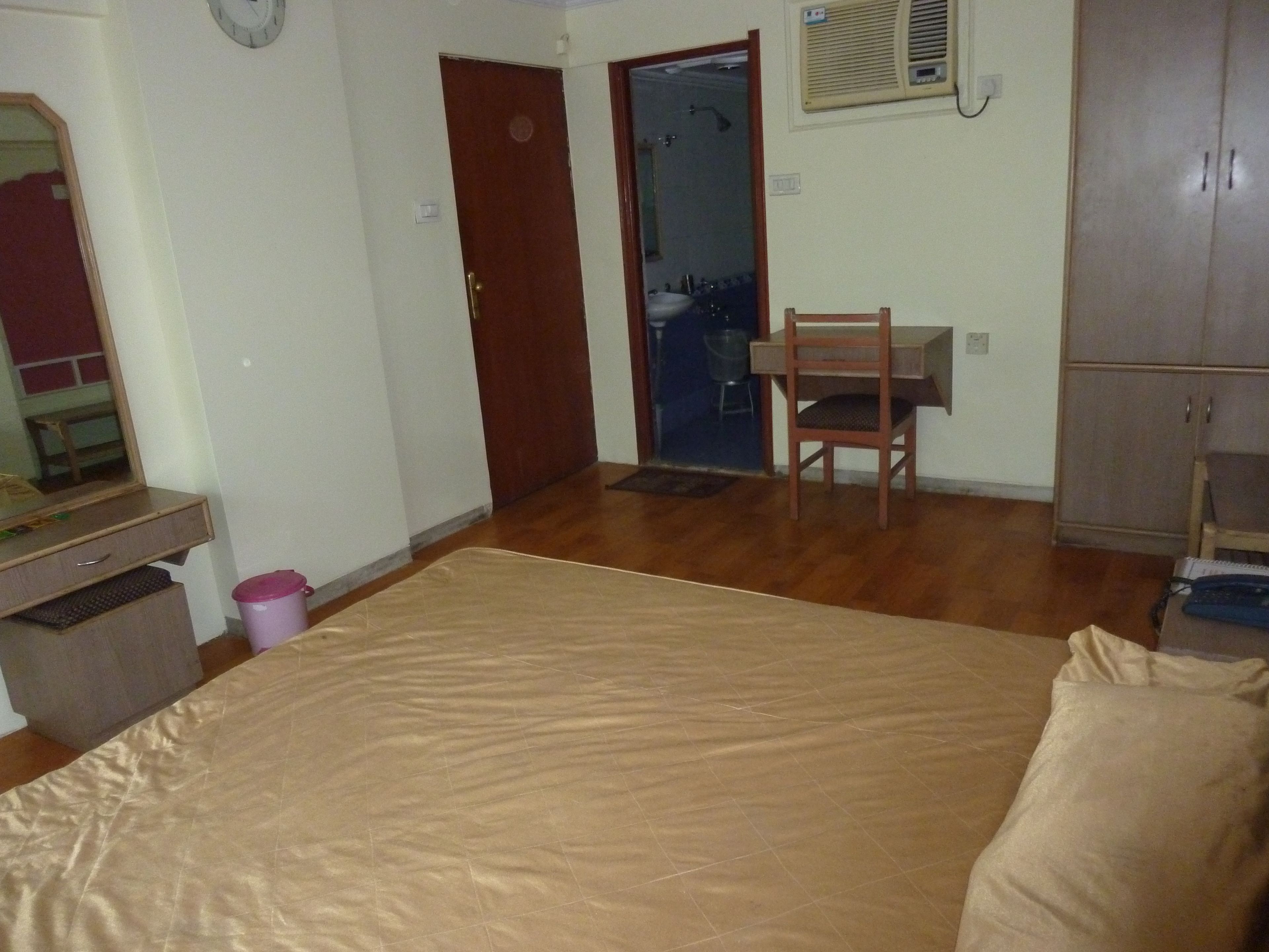Une chambre à coucher avec un lit, un bureau en bois, une chaise, une armoire et une salle de bain visible à travers une porte ouverte.