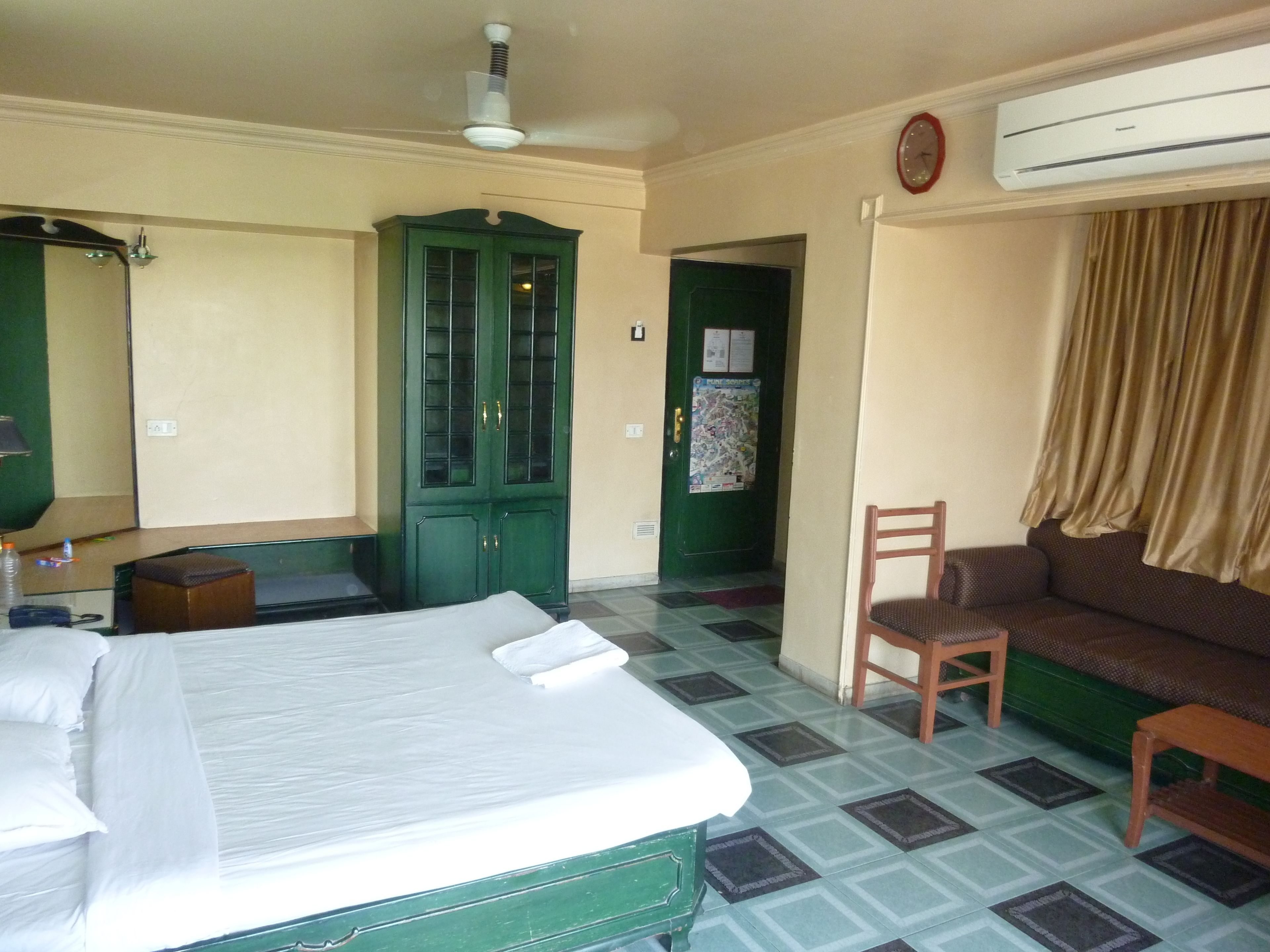 Una habitación de hotel con una cama, un armario verde, una silla de madera, un sofá y un equipo de aire acondicionado.