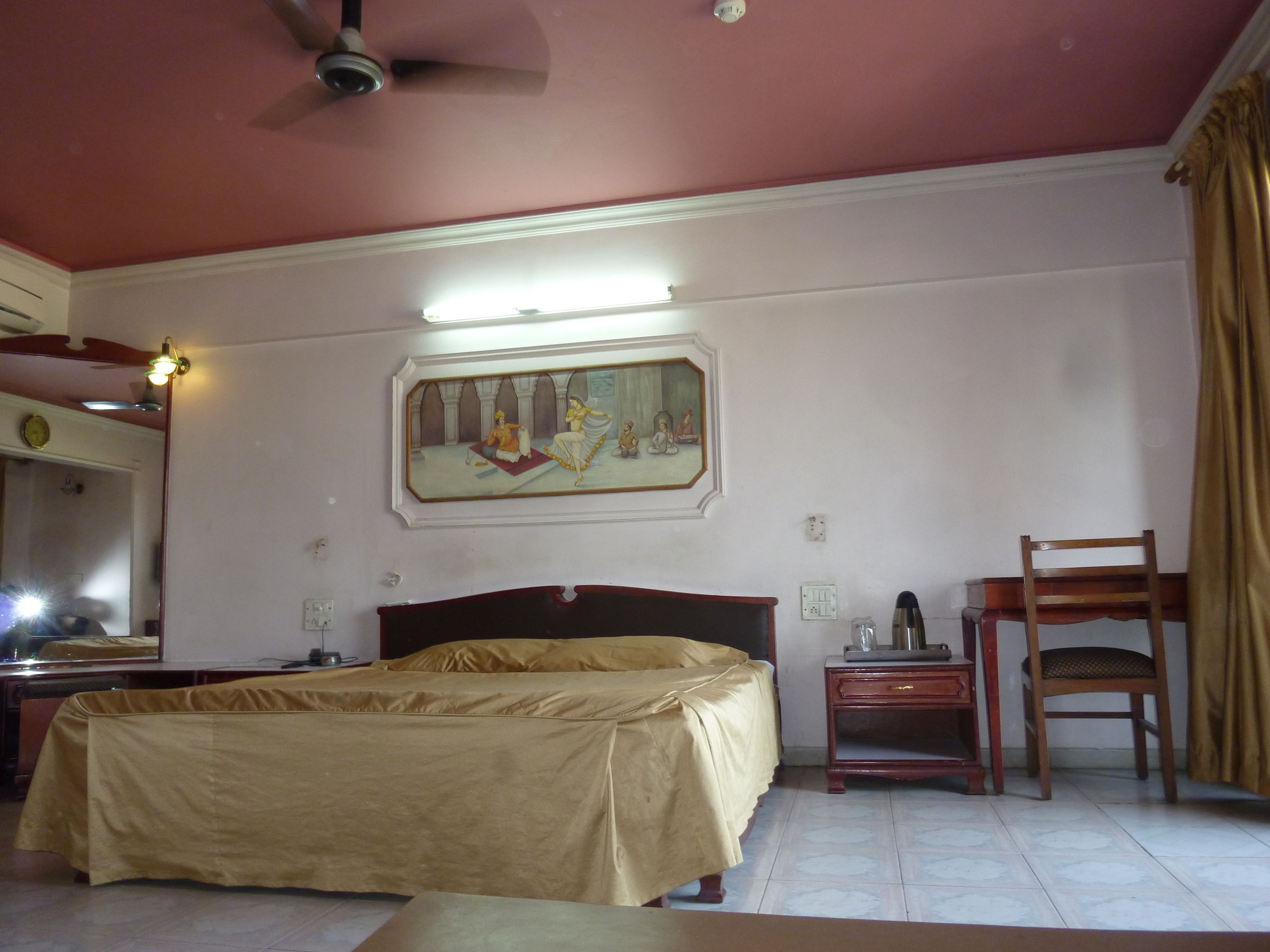 Una habitación de hotel con una cama, una mesita de noche, una silla, un ventilador de techo y un cuadro en la pared.