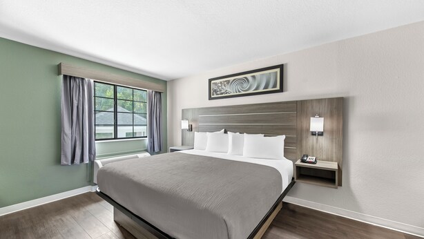 Ein Hotelzimmer mit einem großen Bett, einem Fenster mit Vorhängen, einem Nachttisch mit Lampe und einem an der Wand hängenden Kunstwerk.
