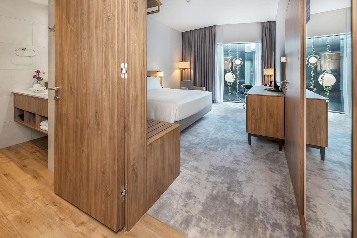 A modern hotel room with a wooden wardrobe, a bed, a desk, and a large window with curtains.
