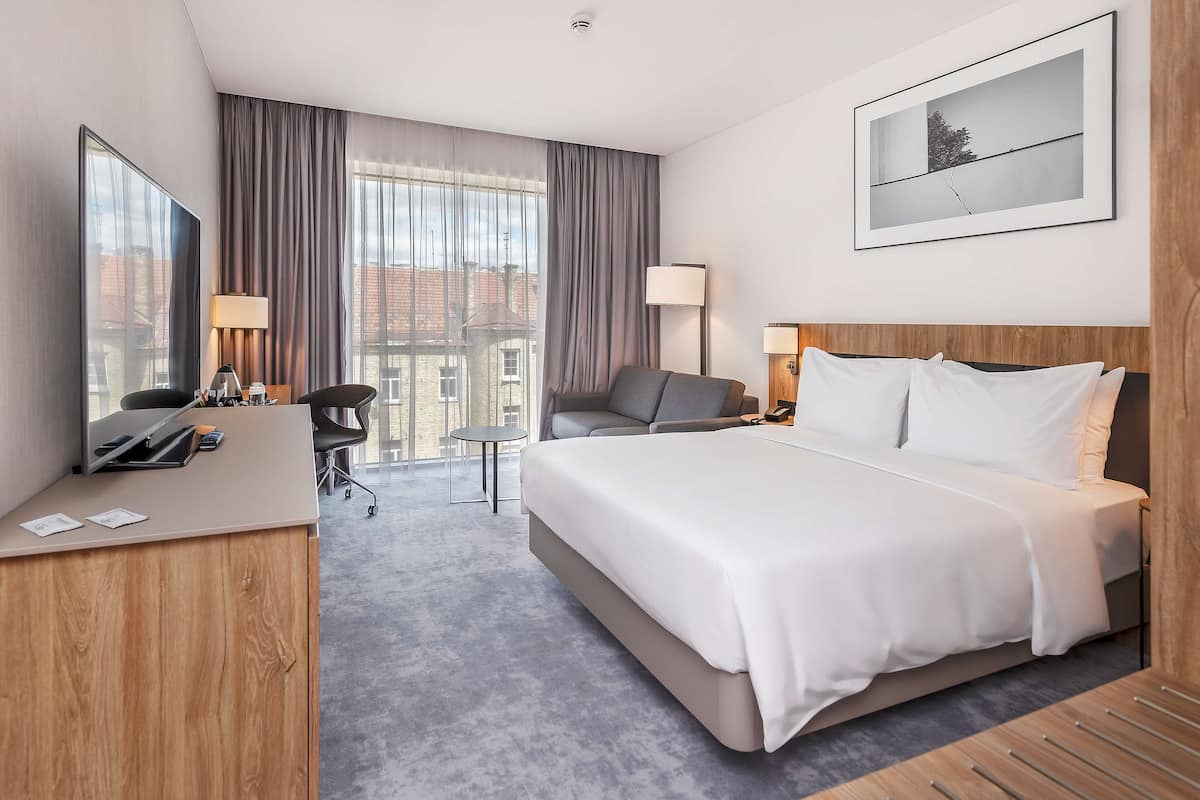 A modern hotel room with a large bed, a desk, a chair, and a view of the city through large windows.