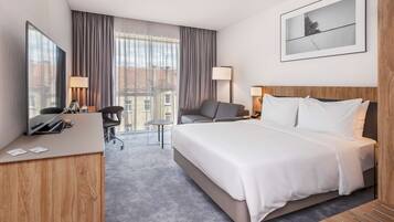 Une chambre d’hôtel moderne dotée d’un grand lit, d’un bureau, d’une chaise et offrant une vue sur la ville grâce à de grandes fenêtres.