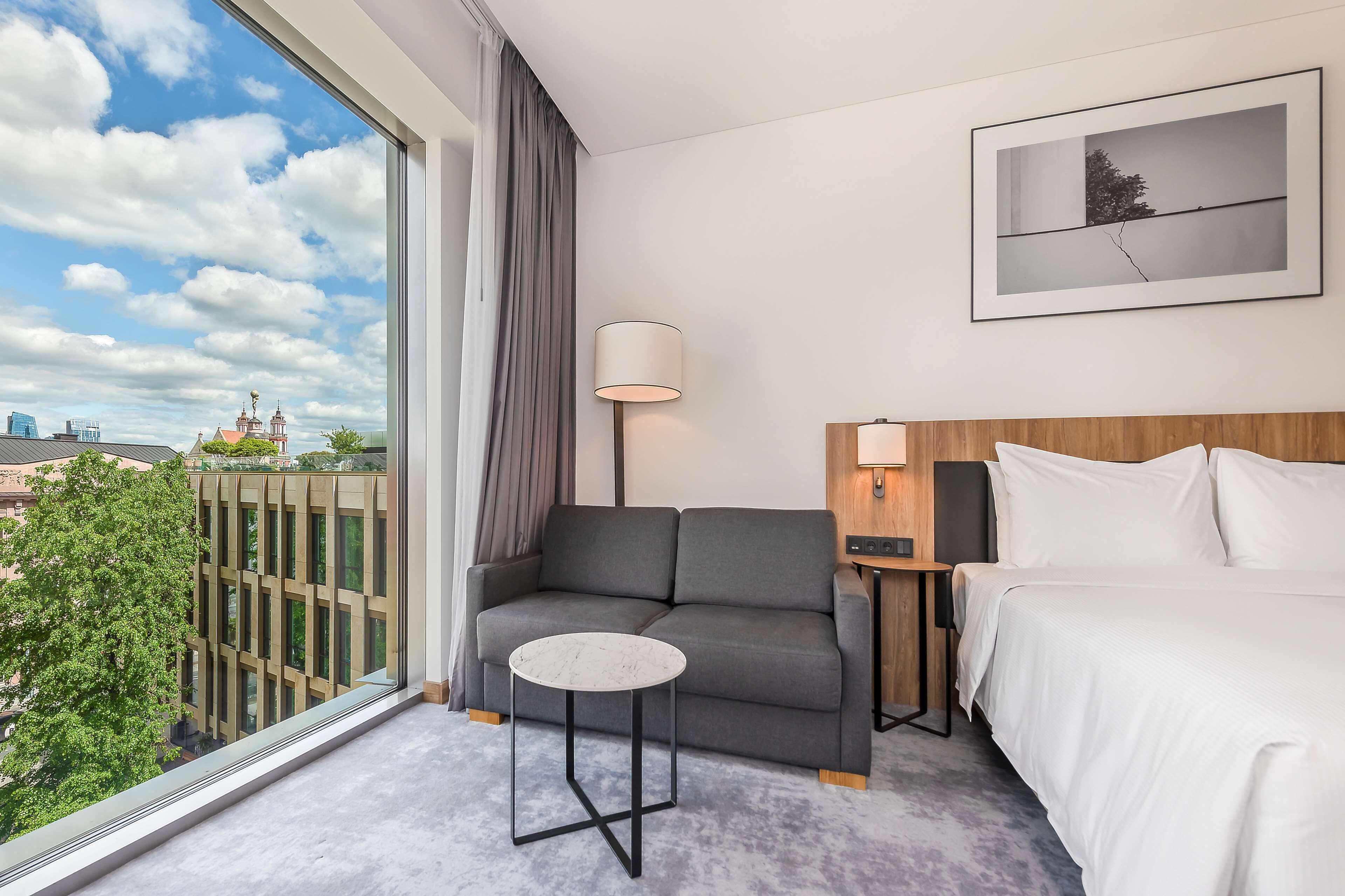 A hotel room with a sofa, a small table, a bed, a lamp, and a framed picture on the wall.