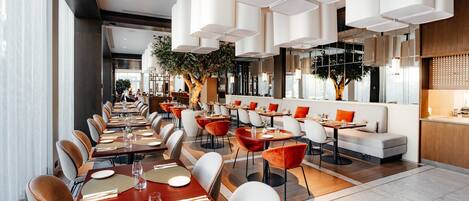 A modern restaurant with long communal tables, individual booths, and a mix of red and grey chairs.
