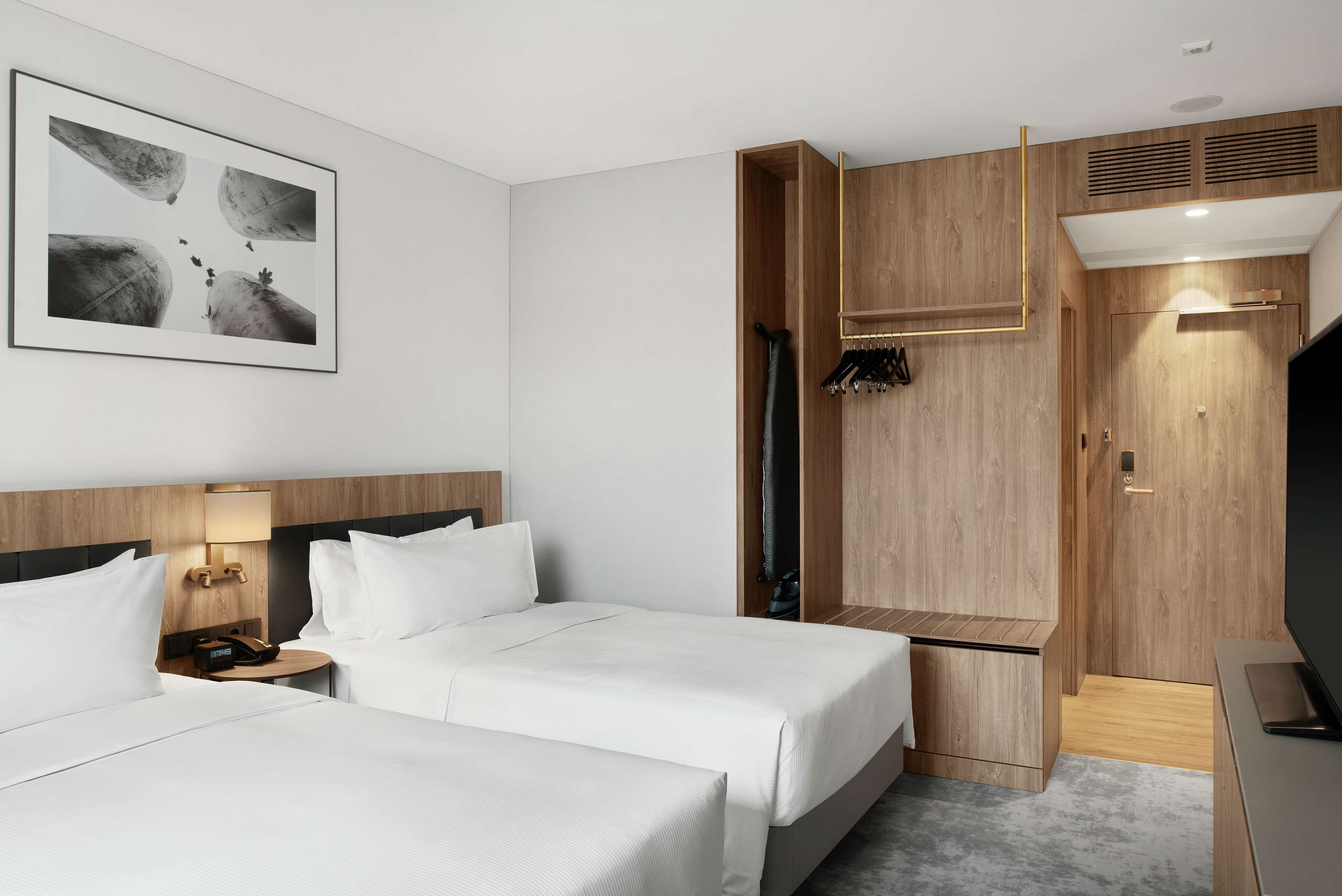 Ein Hotelzimmer mit zwei Betten, einem Holzschrank, einem Kleiderständer und einem gerahmten Kunstwerk an der Wand.