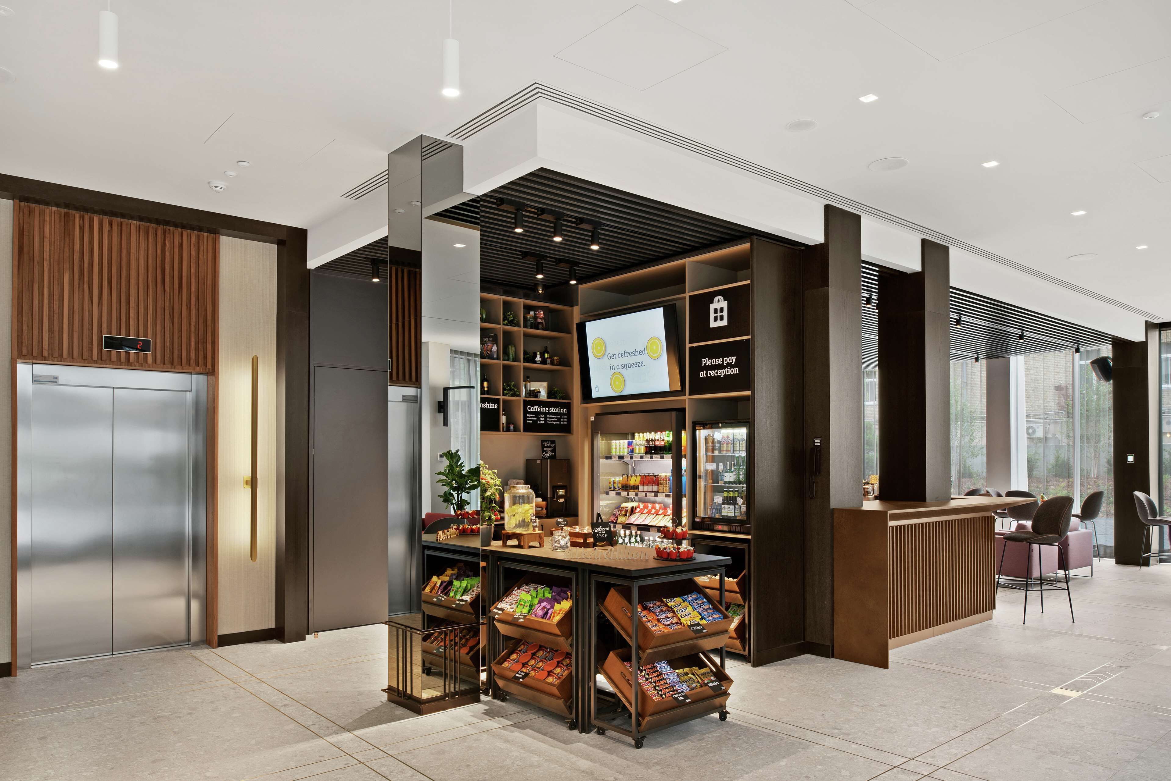 A modern coffee station with a digital display, a refrigerated display case, and a seating area with chairs.
