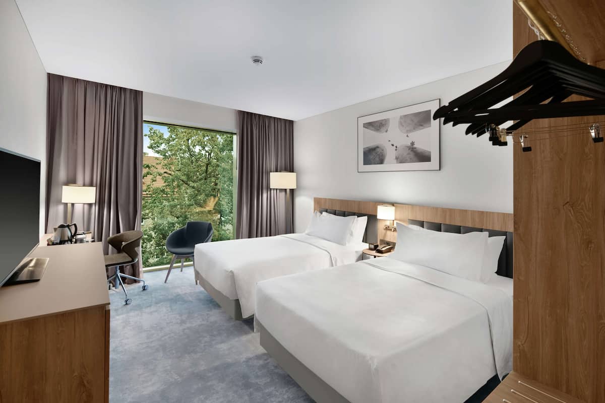 A hotel room with two beds, a desk, a chair, a TV, and a window with curtains.