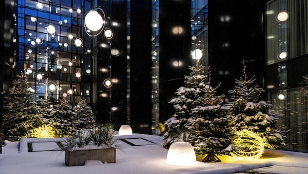 Un espace extérieur enneigé, agrémenté de sphères lumineuses et d’arbres de Noël, situé devant un bâtiment moderne aux fenêtres en verre.