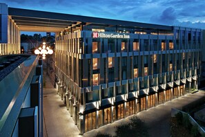 Un hôtel Hilton Garden Inn moderne, doté d’une façade en verre, de fenêtres illuminées et d’une entrée imposante.