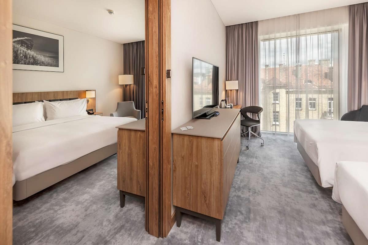 A hotel room with two beds, a desk, a chair, a TV, and a window with a view of buildings.