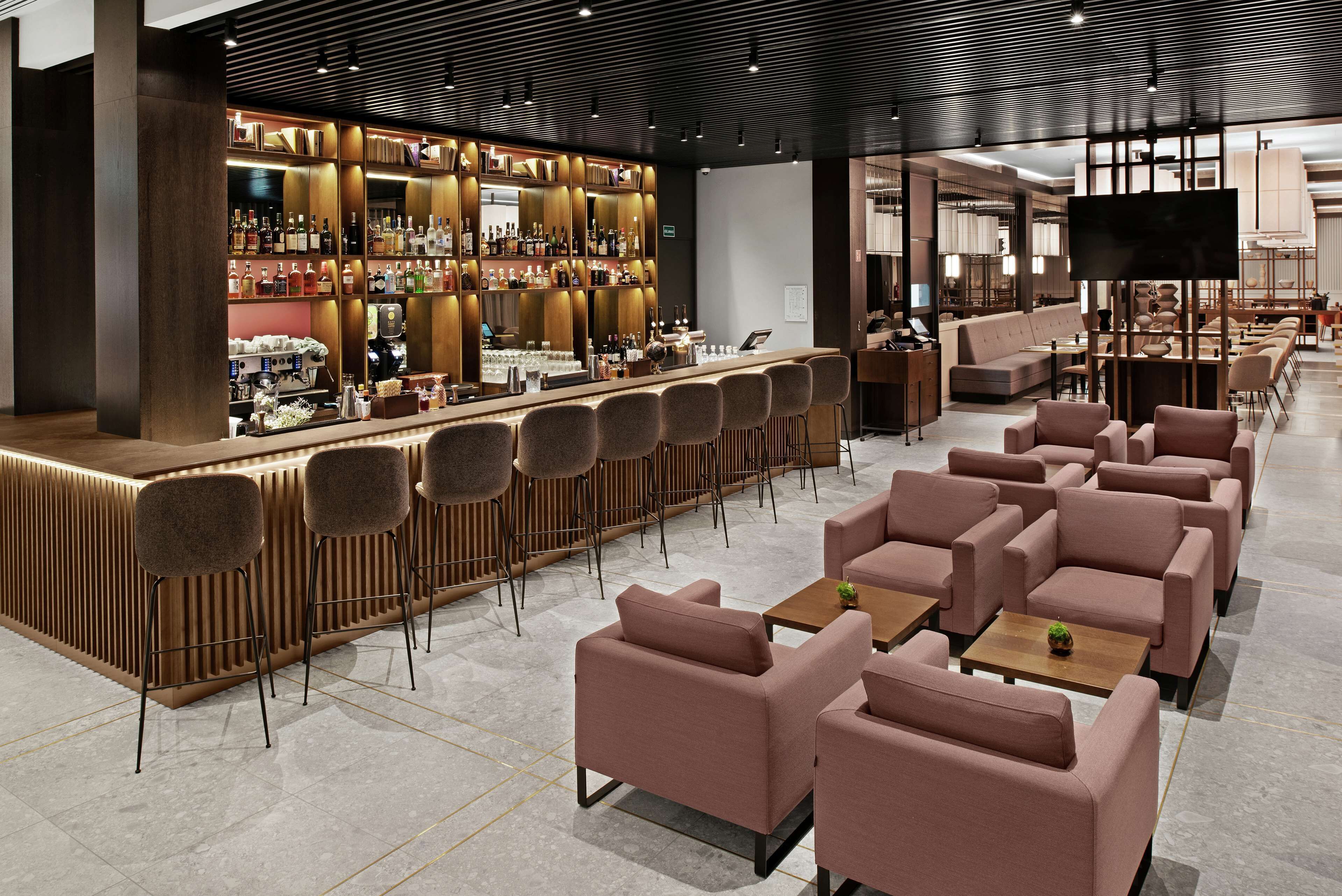 Un área de bar moderna con una barra alargada, varios taburetes y un espacio de estar con sofás y mesas.