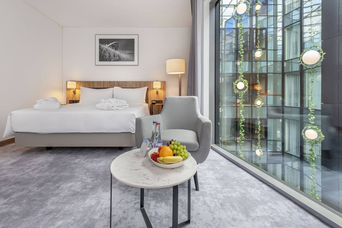 A hotel room with a bed, bedside lamps, a chair, a small table with fruit, and a large window with a view of a building.