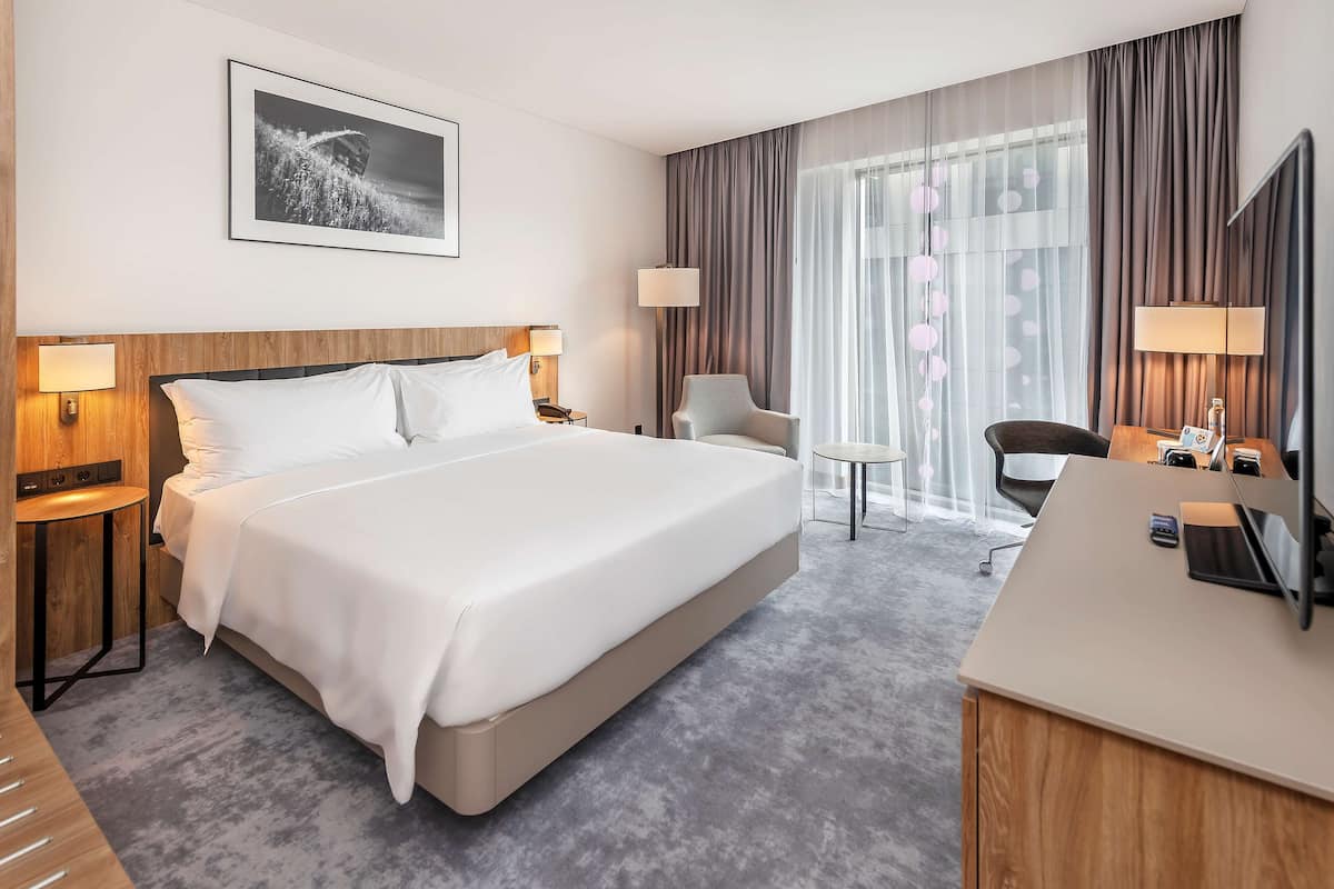 A hotel room with a large bed, bedside tables with lamps, a desk with a chair, and a large window with curtains.