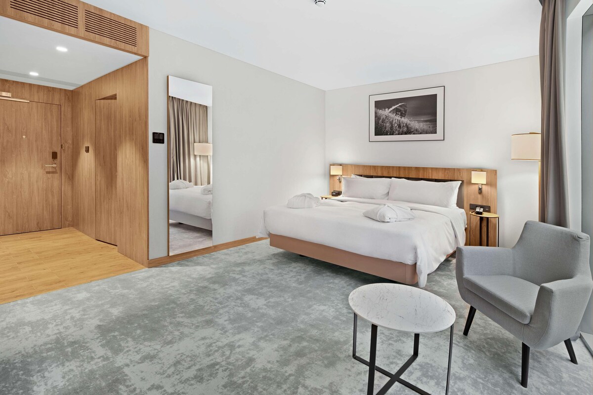 Una habitación de hotel moderna con una cama grande, un sillón gris, una mesita redonda pequeña y un armario de madera.