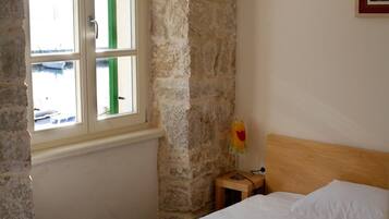 Une pièce avec un lit, une table de chevet, une lampe, une fenêtre avec des volets verts et un mur en pierre.