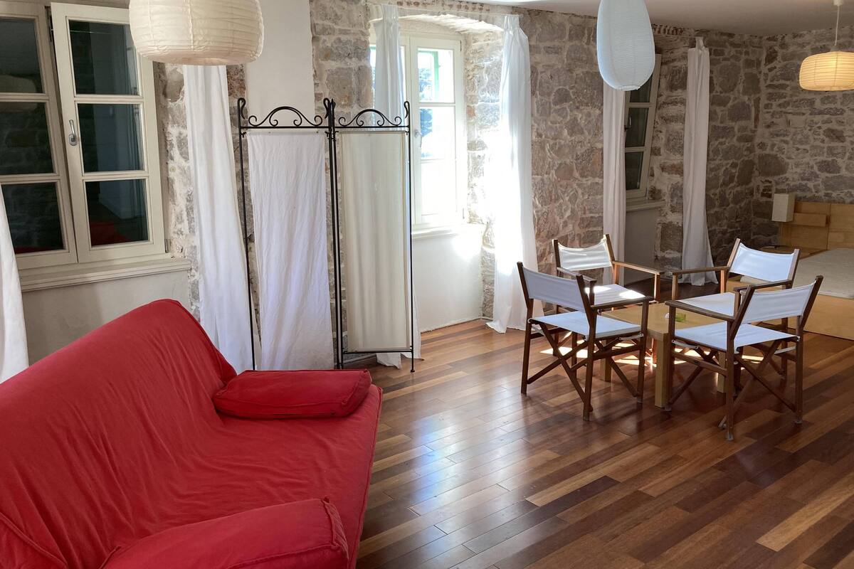 A room with a red sofa, wooden dining table with chairs, stone walls, and a window with curtains.