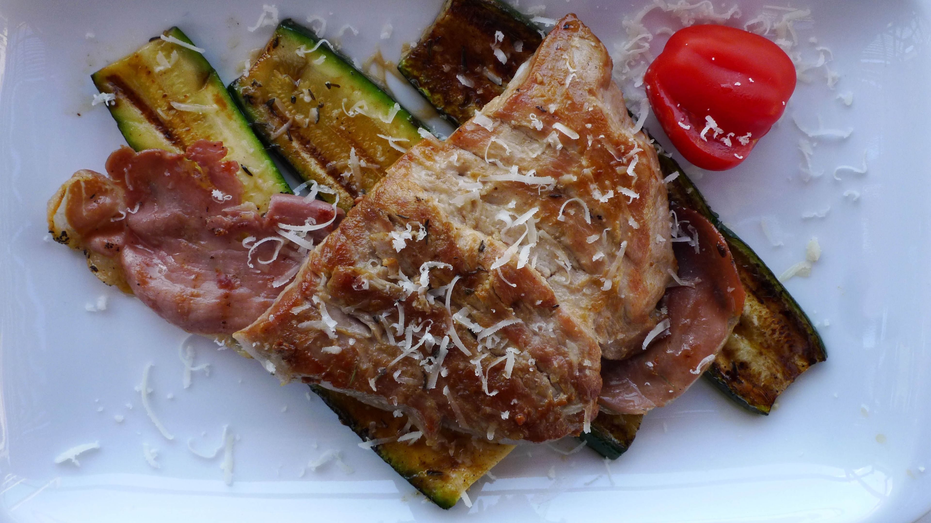 A meal with grilled vegetables, a slice of ham, and a tomato on a white plate.