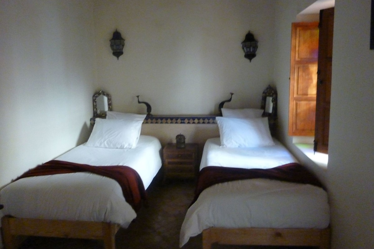 Una habitación con dos camas, una mesita, lámparas fijadas a la pared y una puerta de madera.